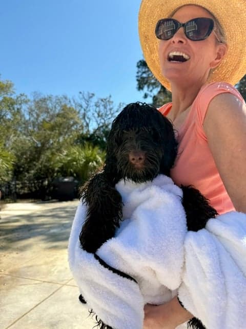 A woman coming back from a beach day with their black Australian Labradoodle wrapped up in a beach towel and they're both smiling.